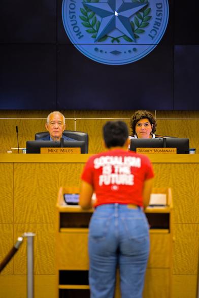 Center-frame is a person speaking at a podium wearing a red shirt emblazoned with the phrase "socialism is the future". They are out of focus.

Behind them sit two people at an elevated seating area: Audrey Momanaee and Mike Miles, the latter being the current state-placed superintendent of HISD. They look on at our speaker, with Miles seemingly uninterested in anything they have to say.