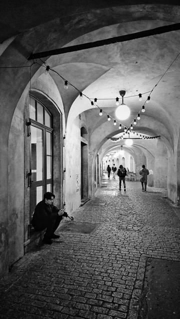The image is a black and white portrait-orientated photograph of a covered walkway with a cobblestone ground and arched ceilings. String lights with exposed bulbs are suspended from the ceiling throughout the walkway. A large, round light fixture is also visible hanging from the ceiling further down the passage. On the left side of the walkway, there is a building wall with several arched doorways and windows. In the foreground, a person is sitting on a ledge next to a doorway. Further down the walkway, several other individuals are walking. The perspective is from the near end of the walkway looking down its length.