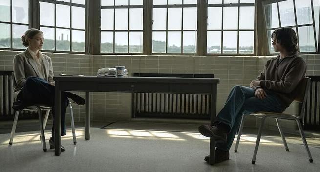 Amanda Seyfried and Tom Holland sit in defensive positions across the length of a table. A wall of windows is behind them.