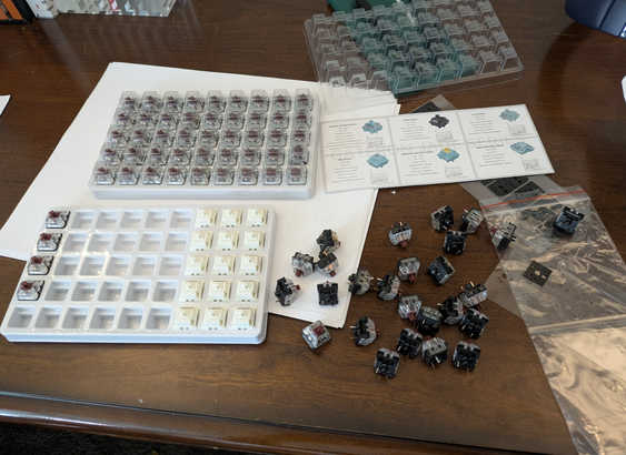Messy wood desk with a open box of gateron smoothie switches and a pile of kaliah copper switches next to it.