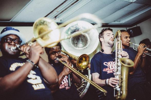Deux joueurs de trombone, un saxophoniste et un trompettiste soufflent à plein poumon sur une petite scène. Le flash fait briller les cuivres.