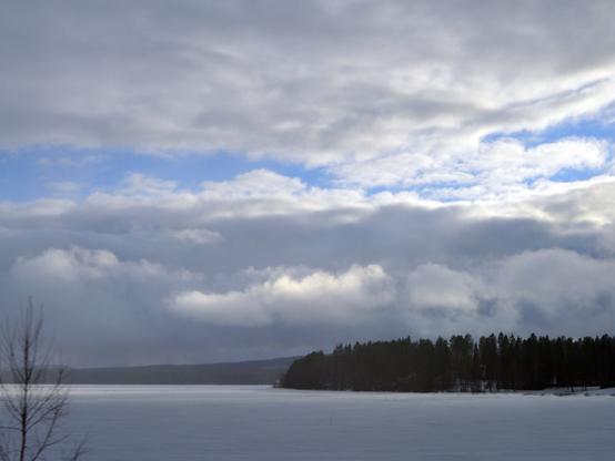 Mostly clouded sky with patches of blue, over an icy lake/river, with a distant peninsula/island with a trees.
