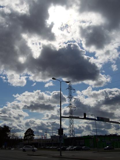 Clouds partially obscure sunlight over a street with traffic lights. Commercial buildings can be seen across the street. A high voltage power pole can be seen towering behind them.