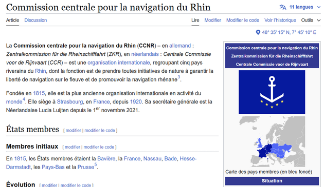 Capture d'écran de l'article Wikipédia en français sur la Commission centrale pour la navigation du Rhin (CCNR). L'article présente la CCNR comme une organisation internationale regroupant cinq pays riverains du Rhin, ayant pour mission de garantir la liberté de navigation sur le fleuve et de promouvoir la navigation rhénane. Fondée en 1815, elle est la plus ancienne organisation internationale encore en activité. Son siège est situé à Strasbourg, en France, depuis 1920. L'image inclut également une carte des pays membres en bleu foncé et un drapeau avec une ancre entourée d'étoiles.