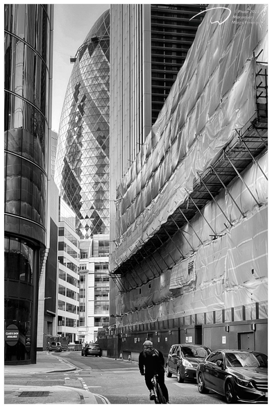 See through in one if the back streets on Tower Hill, London. The famous gherkin tower is visible in between other buildings, a cyclist rides in the street.