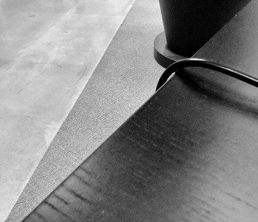 Abstract black and white image showing a corner of a desk, a cable, a rug, and a big flowerpot, with textures and shadows creating visual depth.