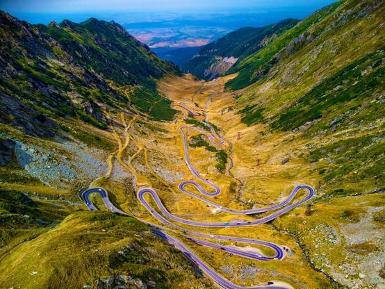 Aerial view of the Transfăgărășan road in Romania, showcasing its winding path through lush green mountains with sharp turns and a scenic valley in the background.