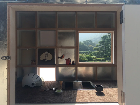 Blick in das Innere des Wagens. Auf Hüfthöhe ist eine Tatami-Matte als Boden ausgelegt. Darin eingelassen ist eine quadratische schwarze Vertiefung. Im Vordergrund steht ein runder goldener Teller, auf dem eine Teetasse und Teezubehör stehen. Rechts im Hintergrund ist der Blick durch das geöffnete Fenster.