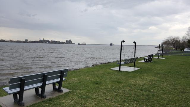 Quelques bancs en face du fleuve, des usines sont situées en arrière plan avec deux bateaux qui se croisent.
