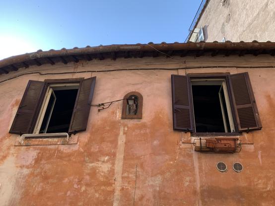 Blick auf die rötliche Fassade eines alten Hauses. Links und rechts ein Fenster mit geöffneten Fenstern und braunen Fensterläden. In der Mitte zwischen den Fenstern eine kleine Nische mit der Figur eines Heiligen. Davor eine kleine Laterne an einem Ausleger, der zwischen dem linken Fenster und der Nische an der Fassade befestigt ist, aber vor die Nische abgedrückt wurde. Die Stromzufuhr erfolgt mit einem Draht und Klemme, die aus dem Fenster geführt werden.