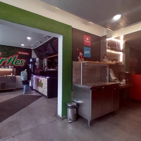 Inside the pizzeria seating area. Order counter (with customers), beverage fridge and salad desk can be seen on the adjacent room to the left. An empty counter and a table with coffee maker, cups and napkins can be seen on the right side. Above the table is a shelf with utensils, and TMNT-themed figurines and painting on the shelf above it.
