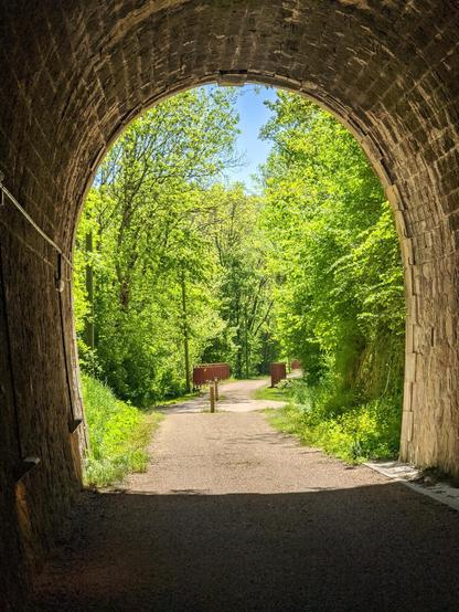 photo prise de l'intérieur d'un tunnel  sur une voie verte (ancienne voie ferrée), en regardant vers la sortie. On voie beaucoup d'arbres bien vert, au loin les garde-corps d'un pont