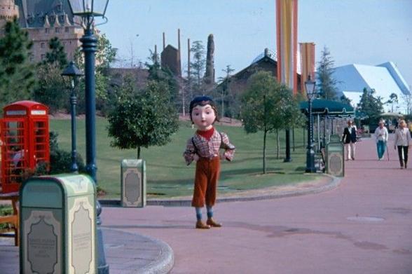 A large, cartoonish character figure stands on a path in a park, surrounded by greenery and trees. In the background, a red phone booth and people walking can be seen, along with colorful flags and a building structure.