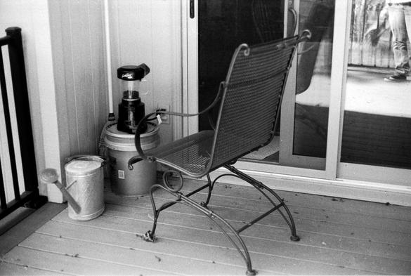 A wrought iron deck chair is pulled up to a corner in which a small coffee roaster sits on top of a gallon bucket.