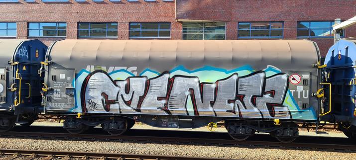 A side shot of a brown bulk freight train covered in a single piece of graffiti. It consists of of grey letters with white striping and highlights with black sides with red striping. It reads cmenents. This against a lime green with blue outline, possibly of an earlier piece.