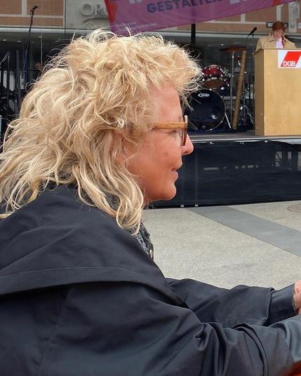 Beate Müller-Gemmeke bei einer 1.-Mai-Kundgebung des DGB. Im Vordergrund im Profil, im Hintergrund eine Rednerin am DGB-Rednerpult auf einer Bühne.