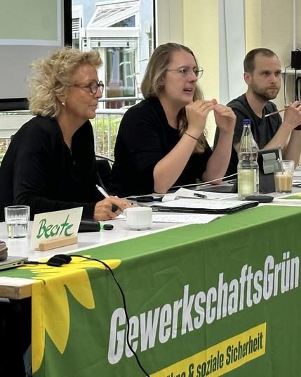 Beate Müller-Gemmeke bei einer Sitzung von GewerkschaftsGrün. Sie sitzt neben anderen Teilnehmenden hinter einem Tisch mit grünem Banner und diskutiert konzentriert.