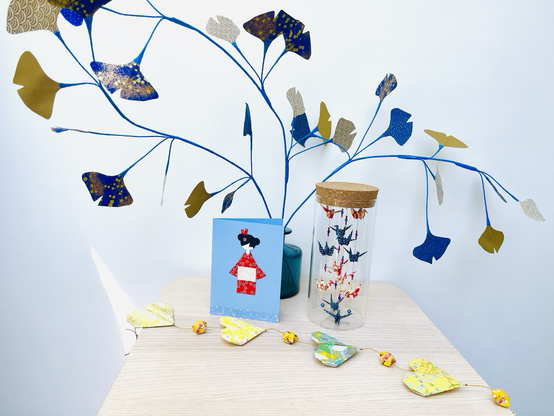 Sur une table blanche une volière bleue et rouge, une carte, une guirlande de cœurs jaunes et un bouquet de gingko le tout en origami
