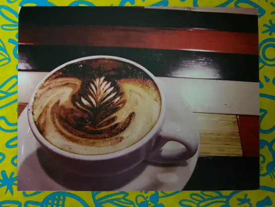 Postcard showing a cup of coffee decorated with latte art in a white cup on a white saucer on a wooden table.
The table is mad of narrow strips of different colour wood.