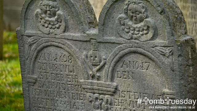 Zwei verwitterte, nebeneinanderstehende historische Grabsteine aus Sandstein mit kunstvoller Reliefgestaltung. Beide Steine tragen im oberen Bereich ein rundliches Engelsgesicht mit stilisiertem, lockigem Haar und angedeuteten Flügeln unter dem Kopf – vermutlich Putten oder Todesengel. Zwischen den Grabplatten ist ein kleiner Totenschädel mit gekreuzten Knochen und einer umgedrehten Sanduhr zu sehen – ein klassisches Vanitas-Symbol für die Vergänglichkeit des Lebens. Die eingemeißelten Inschriften sind teils noch gut lesbar und datieren auf die Jahre 1698 und 17XX.