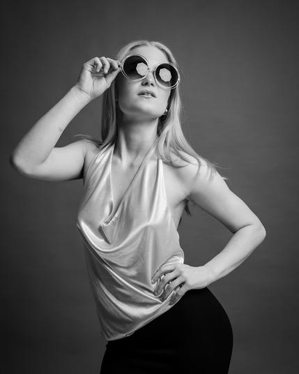 This black and white image features a person striking a pose, wearing large round sunglasses and a sleeveless, draped top. They have long hair and are standing against a plain background. One hand is on their hip, and the other is adjusting the sunglasses.
