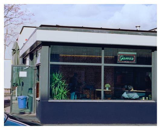 Color photograph of a gas station diner in Vienna. The photo is taken from the outside and shows the large window front with a neon sign that reads “Games” and under which a person is sitting leaning against the window pane. A blue garbage can and a closed parasol can be seen through the open door on the left-hand side of the picture. Two other people in the diner are not clearly recognizable and are standing in the shadows.

Translated with DeepL.com (free version)