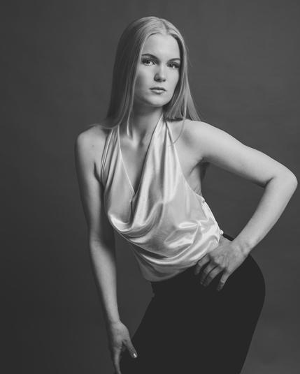 The image is a black and white portrait of a person posing. They are wearing a sleeveless top and dark pants. The photograph has a studio setting with a simple backdrop.