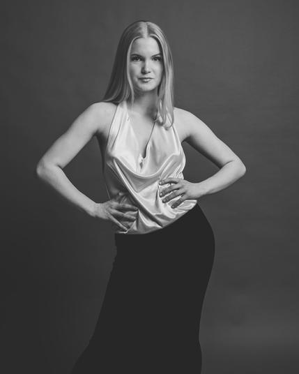 This image is a black and white portrait of a person posing in a studio setting. They are wearing a sleeveless, shiny top and a long, dark skirt. The person has long hair and is standing with their hands on their hips.