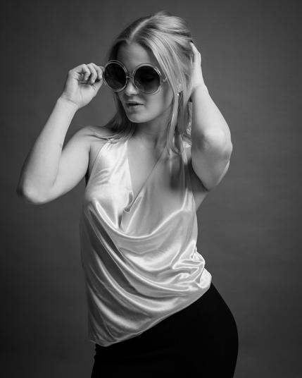 The image is a black and white portrait of a person wearing large round sunglasses and a sleeveless top with a draped neckline. The person's hair is styled back, and they are posing with one hand touching the sunglasses and the other hand in their hair. The background is plain, emphasizing the subject.