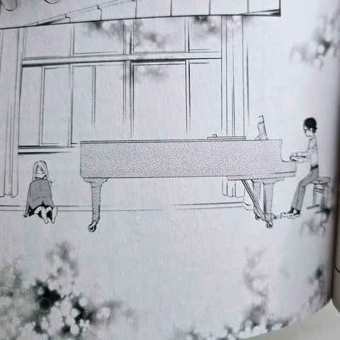 Photograph of a black and white manga page from the interior of Your Lie in April, Volume 3. The panel shows a teenage boy playing the piano on a large, grand piano with a light haired teenage girl sitting on the ground asleep beneath a large series of windows. The edges are panel are stylized to appear as though cherry blossoms are falling or surrounding the panel.