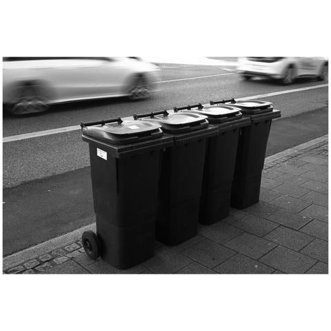 Picture of four wheelie bins in a row on a sidewalk. In the background a road with two cars partly visible. These are quite blurred due to slow shutter speed.