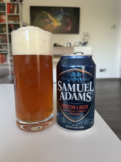 Das Bild zeigt eine Samuel Adams Boston Lager-Can, die auf einem Tisch steht. Die Dose ist dunkelblau mit einem silbernen Deckel und hat ein Emblem mit einem Mann, der ein Bierglas hält. Der Text auf der Dose lautet "Samuel Adams" in großen weißen Buchstaben, gefolgt von "Brewer Patriot" in kleineren blauen Buchstaben. Darunter steht "Cheers to You" in weißer Schrift, gefolgt von "Boston Lager" in roter Schrift. Weiter unten steht "Distinctively Complex & Balanced" in weißer Schrift. Links neben der Dose ist ein Glas Bier zu sehen.