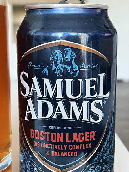 Das Bild zeigt eine Flasche Samuel Adams Boston Lager neben einem gefüllten Glas Bier. Die Flasche ist dunkelblau mit einem silbernen Deckel und hat ein Etikett mit der Aufschrift "Samuel Adams Boston Lager" in großen weißen Buchstaben. Darunter steht "Distinctively Complex & Balanced" in roter Schrift. Das Etikett zeigt auch eine Illustration eines Mannes, der ein Glas Bier hält, und die Angaben "12 FL. OZ. LAGER | 5.0% ALC./VOL. | 30 IBUs". Das Glas ist transparent und zeigt das goldbraune Bier mit einer weißen Schaumkrone. Im Hintergrund ist ein gerahmtes Kunstwerk und ein Bücherregal zu sehen, was auf eine gemütliche, möglicherweise häusliche Umgebung hinweist.