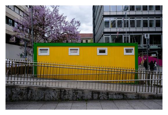 Foto im Querformat. Großstadt, vor der Kamera verläuft ein leicht ansteigender Fußweg von rechts nach links, auf beiden Seiten Geländer. Im Hintergrund sind hohe Häuser. Direkt hinter dem Fußweg ist ein knallgelber Container. Oben sind vier kleine Fenster. Der Container hat einen grünen Rand. Hinter dem Container steht ein Baum mit fliederfarbenen Blüten. Gelb, grün und Flieder stechen heraus, aus der sonst grauen Farbgebung.