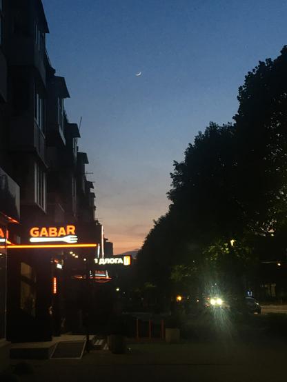 A nighttime street scene in Lutsk, Ukraine, under a dark blue sky with a faint crescent moon. On the left, a multi-story building with a brightly lit orange sign that reads "GABAR" is visible. Across the street, silhouetted trees line the right side of the frame. Streetlights illuminate the sidewalk and road, and the headlights of a car are visible in the distance, creating bright streaks of light. Another illuminated sign with Cyrillic text is partially visible further down the street. The overall atmosphere is calm and depicts a typical urban evening.