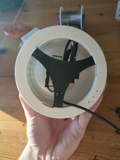 Eine Hand hält ein großes weißes rundes Teil aus Plastik. Darin ein schwarzes Dreieck auf dem ein paar Bauteile auf der Rückseite montiert sind. An der Seite geht ein Kabel raus