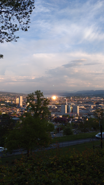 Blick von der Waid auf Zürich und den Zürisee. Kurz vor unteren (nahen) Ende des Sees spiegelt sich die Sonne ineinem grossen Haus mit Glasfront, sodass es aussieht, als würde die Sonne grade im See deutlich vor dem Horizont untergehen.