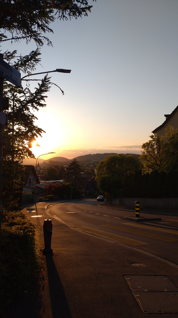 Blick gen Westen auf eine von der Abendsonne golden beleuchtete Strasse einen Hang hinab. Links ein Busch, am Horizont die über Hügeln noch gleissende Sonne, rundherum Abendhimmel von gelb über pink und orange zu mausgrau (Wolken am Horizont) bzw graublau (Himmel darüber).