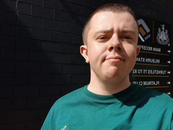 A white transmasc wearing a green t shirt. He has a buzzed head and is stood in front of a black brick wall with a sign that has a Newcastle united logo and and arrow towards reception