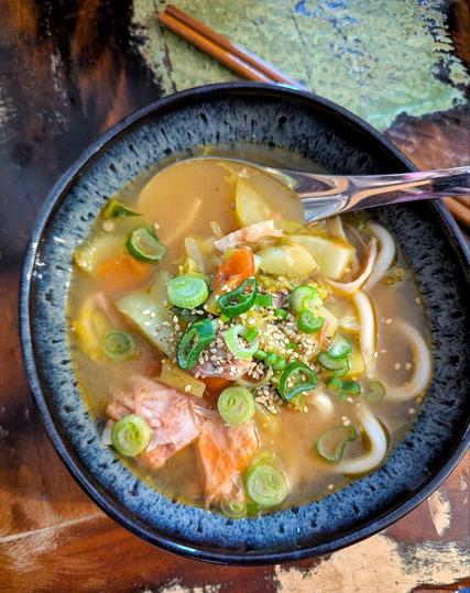 Dunkler Tisch, darauf eine unregelmäßig geformte, grau gemusterte Schale. In der Schale eine gelbliche Brühe mit allerlei  Gemüse, Fleisch und dicken, weißen Nudeln. Darübergestreut ist ungeschälter Sesam und Frühlingszwiebelringe. In der Schale steckt ein metallener asiatischer Suppenlöffel. Neben der Schale liegen Essstäbchen.