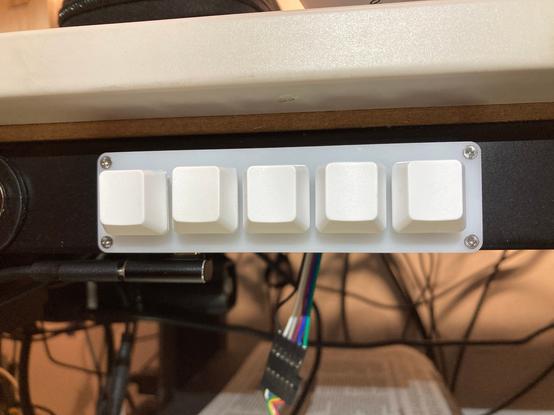 A close up of a 5 key macropad attached to a metal desk frame horizontally. The keypad as a right-angled USB-C adaptor leading a cable off picture to the left. Below it are some dangling wires with a Dupont 6-way connector and coloured ribbon cables which run off out of sight.