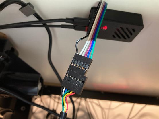 A picture of the underside of a desk. A 3D printed black box about the size of a matchbox is in the background, with two USB-C cables plugged in to the left short edge. A motor housing is blurry off to the left. A red light shows through the black boxes underside and there is a grille to the right. Leading out of the box toward the camera is a 6-way Dupont connector with multi-coloured wired leading out of sight.