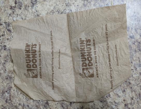 A brown unbleached Dunkin' Donuts napkin on a counter. Both halves of the napkin have DUNKIN' DONUTS in big letters next to the logo above "AMERICA RUNS ON DUNKIN" Below the logo we see "Proudly serving great tasting coffee for over 60 years" and "DunkinDonuts.com"