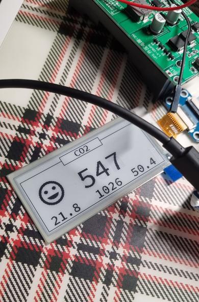 Ein eInk-Display auf dem oben eingerahmt CO2 steht. In der Mitte ist groß die Zahl 547 angezeigt. Daneben ein fröhlicher Smiley. Unten sind die Zahlen 21.8, 1026 und 50.4 angezeigt. Oben im Bild ist eine elektronische Platine zu erkennen.