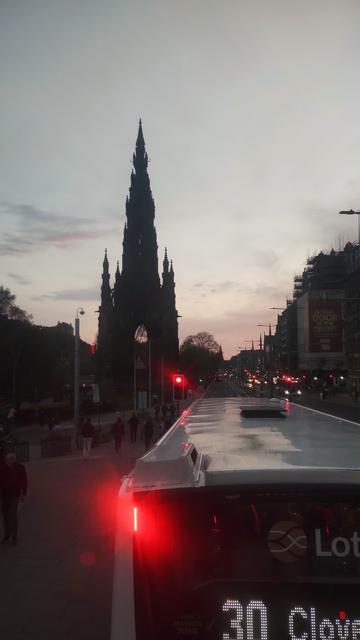 Looking out of the front window of the upper deck of a double decker bus, west along Princes Street at dusk, the Gothic rocket of the Scott Monument on the left, part of Edinburgh Castle on the far left