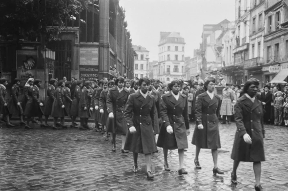 An image of women in military dress walking down cobbled streets. This shows the personnel of the 6888th Central Postal Directory Battalion taking part in a parade in honor of Jeanne d'Arc in Rouen, France, 1945. The predominantly Black Six Triple Eight was the only all-female unit to serve overseas in WWII.