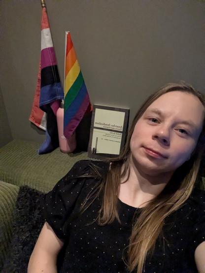 In this candid shot, an individual relaxes on a couch indoors. Two flags stand prominently in the background, along with a water bottle and a book titled Anarchy-Syndicalism.