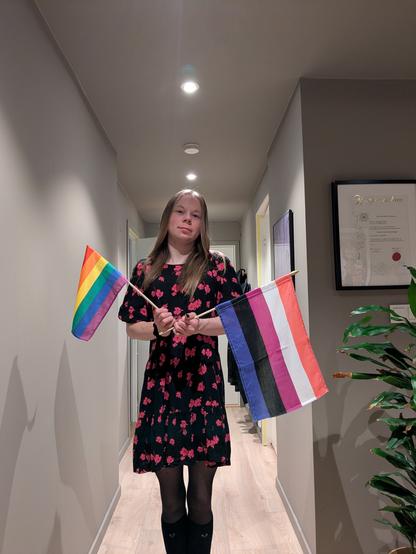 The image features a young person standing in what appears to be a hallway. They are holding a rainbow flag in one hand and a genderfluid pride flag in the other. In the background, there's a framed certificate on the wall and a potted plan.