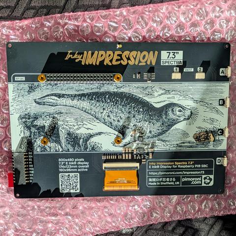 Rear view of the Pimoroni Inky Impression 7.3-inch display, featuring detailed PCB artwork of a seal illustration. Visible are labeled GPIO pins, electronic components, product specs, and the Pimoroni logo, all mounted on the same pink bubble wrap.