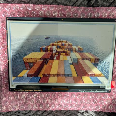 Front view of the Pimoroni Inky Impression 7.3-inch e-ink display, showing a vivid full-color image of a container ship at sea. The screen is mounted on a pink anti-static bubble wrap background and labeled along the bottom edge with product information.